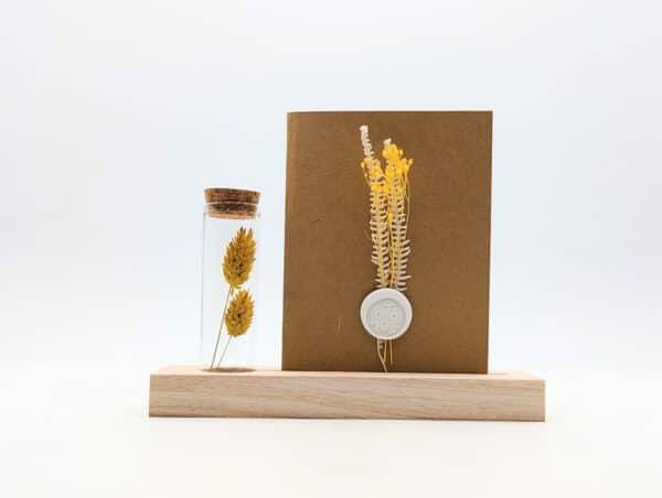 Porte carte en bois avec une éprouvette en fleurs séchées jaunes, carte en kraft brun avec fleurs séchées jaunes et cachet de cire.