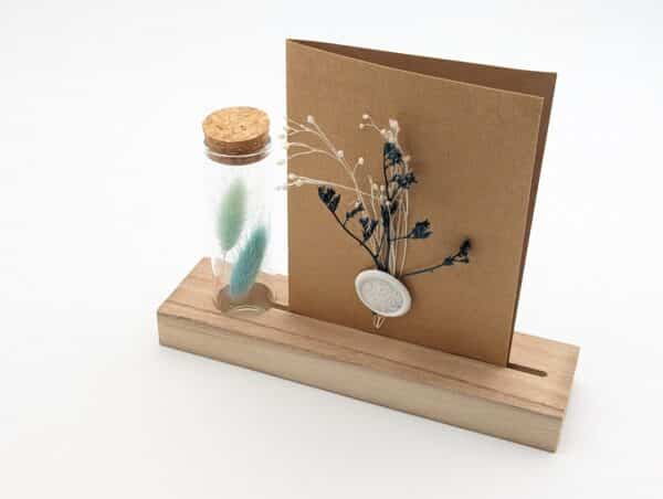 Porte carte en bois avec une éprouvette en fleurs séchées bleues, carte en kraft brun avec fleurs séchées bleues et cachet de cire.