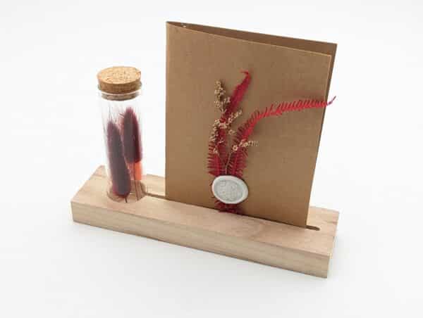 Porte carte en bois avec une éprouvette en fleurs séchées rouges, carte en kraft brun avec fleurs séchées rouges et cachet de cire.
